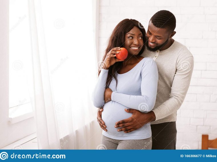 black-couple-love-cuddling-home-next-to-window-healthy-lifestyle-pregnancy-afro-pregnant-wife-eating-apple-husband-166306677